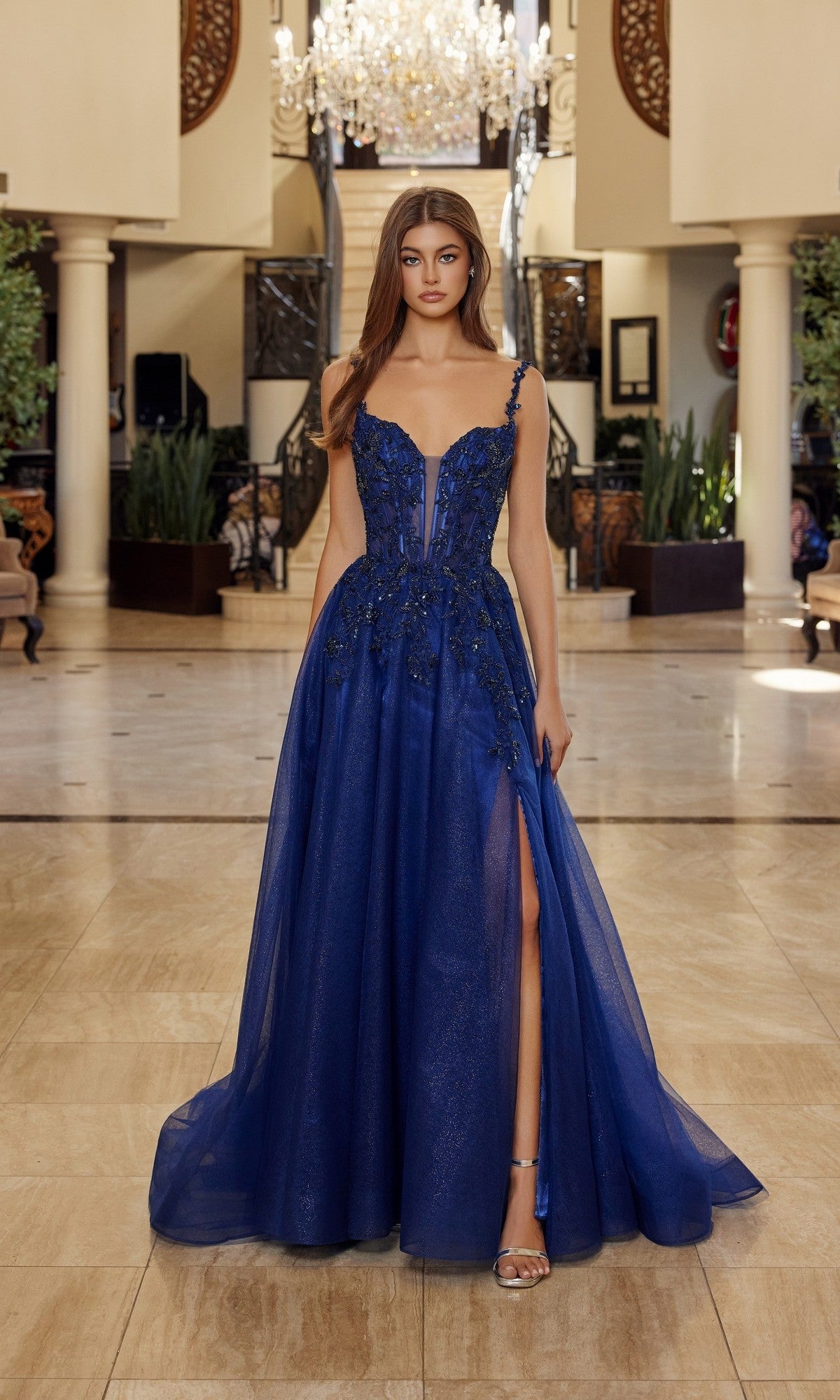 A woman wearing the Nox Anabel R1824 long prom dress, featuring a floral embroidered corset and flowing tulle A-line skirt, stands in an ornate marble interior beneath a sparkling chandelier.